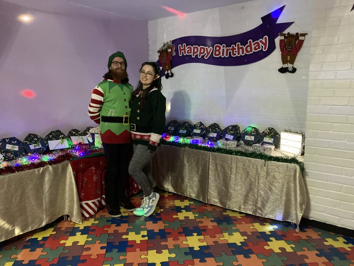 Two volunteers in elf costumes beside a Happy Birthday banner at a RAGS party